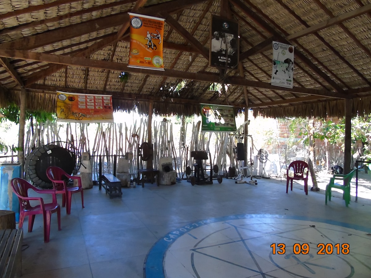An open community space features a thatched roof supported by wooden beams, with concrete flooring. Colorful banners hang above, showcasing local culture and activities. A circle emblem is visible on the ground. Plastic chairs, red and green, are arranged along the perimeter, inviting gatherings.