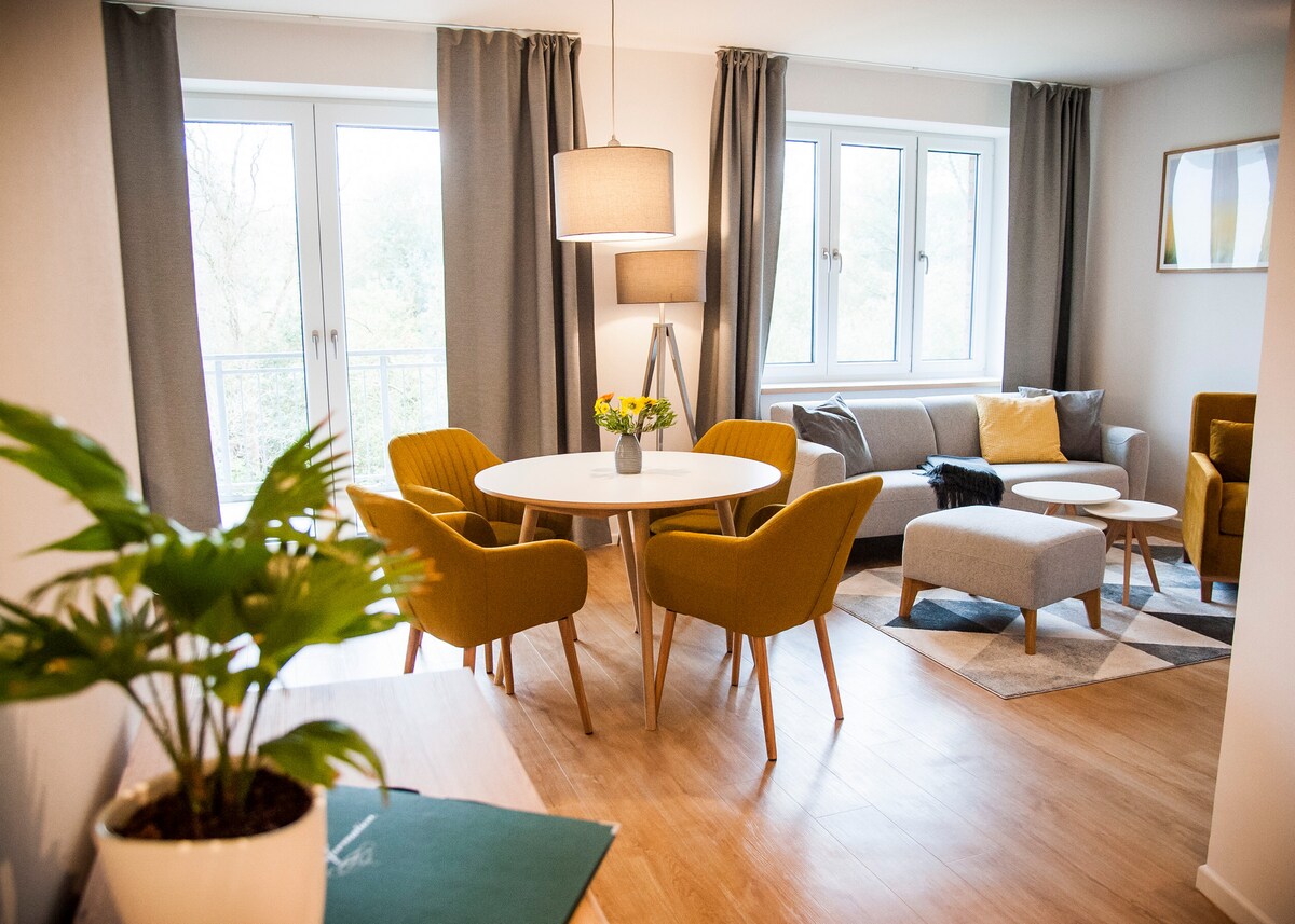 A bright living area is featured with large windows allowing natural light to fill the space. A round dining table is surrounded by four green chairs, while a cozy seating arrangement includes a gray sofa and soft furnishings. A potted plant adds a touch of greenery.