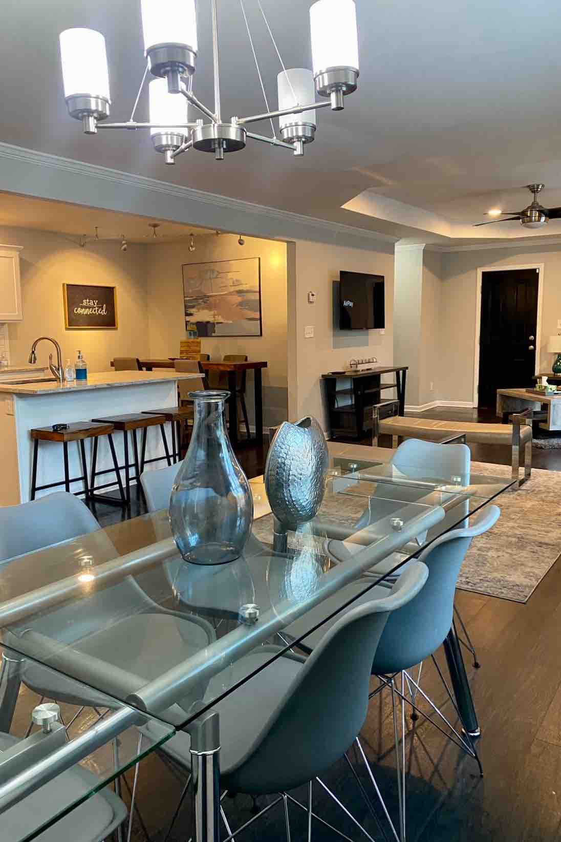 A modern dining area features a sleek glass table surrounded by several chairs. A contemporary chandelier hangs above, illuminating the space. In the background, an open kitchen is visible, showcasing a bar with stools and a view of the cozy living area.