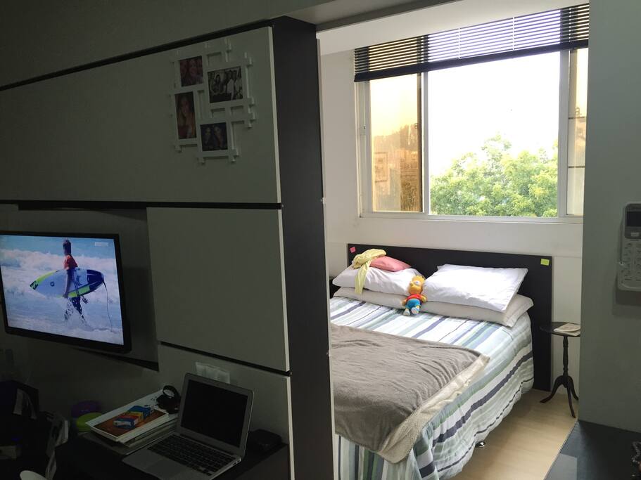 Foto do quarto com cama de casal e ar condicionado e parte da sala com televisão e tv a cabo.