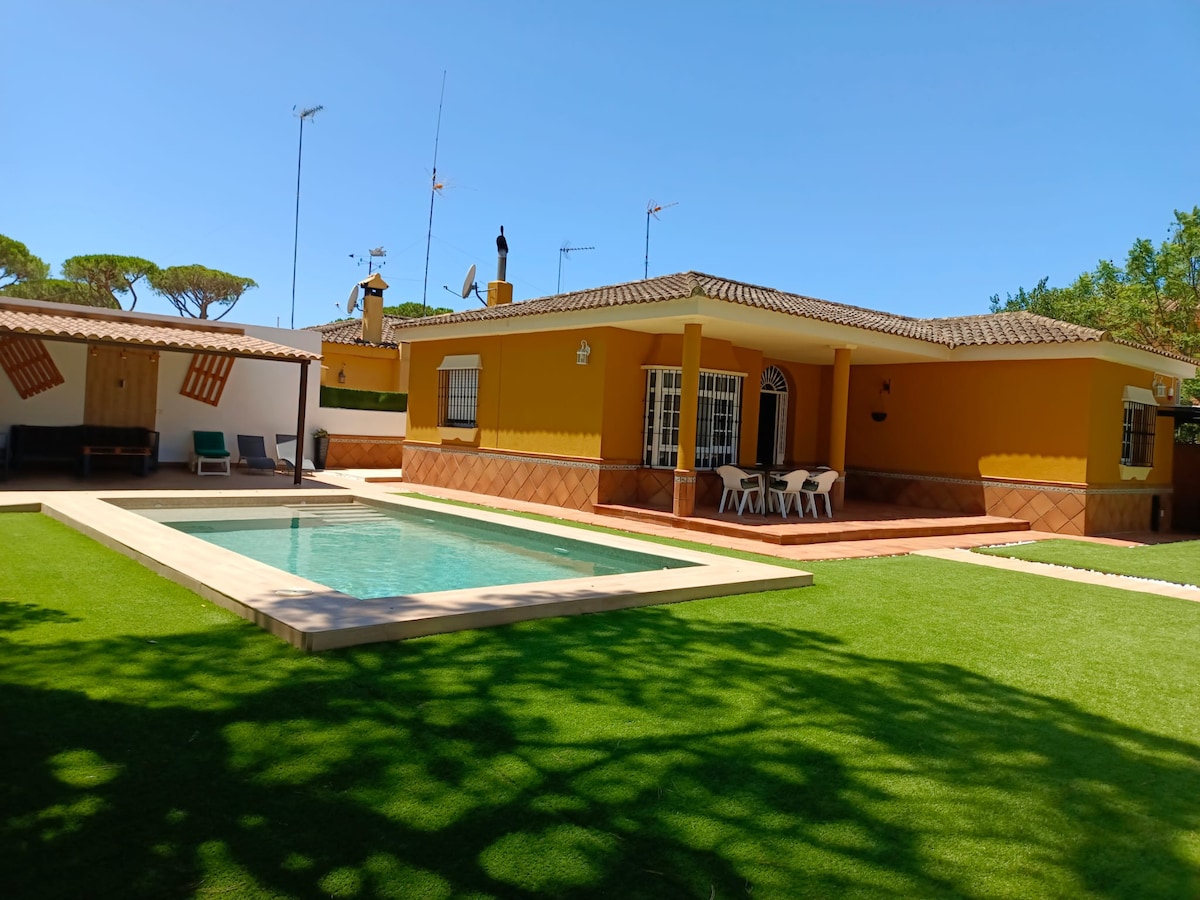 A villa with a bright yellow façade features a well-kept garden with artificial grass. A private pool is centered in the space, accompanied by a covered terrace outlined by white patio chairs. The area is shaded by surrounding trees, providing a tranquil setting.