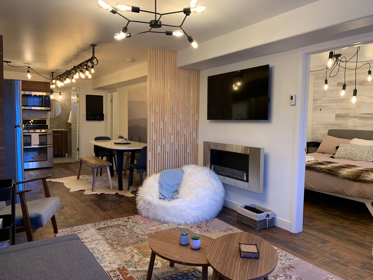 A stylish living area is observed, featuring a cozy, fluffy chair and a modern coffee table with small plants. A dining table sits nearby, illuminated by string lights. The space includes a mounted TV and an electric fireplace, with a view into the bedroom area beyond.