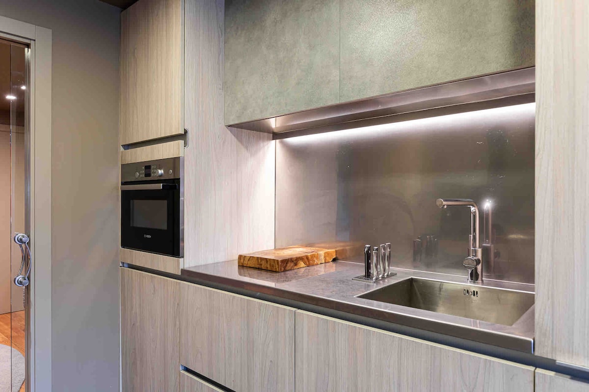 A modern kitchen area features sleek wooden cabinetry and a stainless-steel backsplash. A contemporary sink with a polished faucet is situated next to a wooden cutting board. Built-in appliances, including an oven, are integrated into the cabinetry, enhancing functionality.
