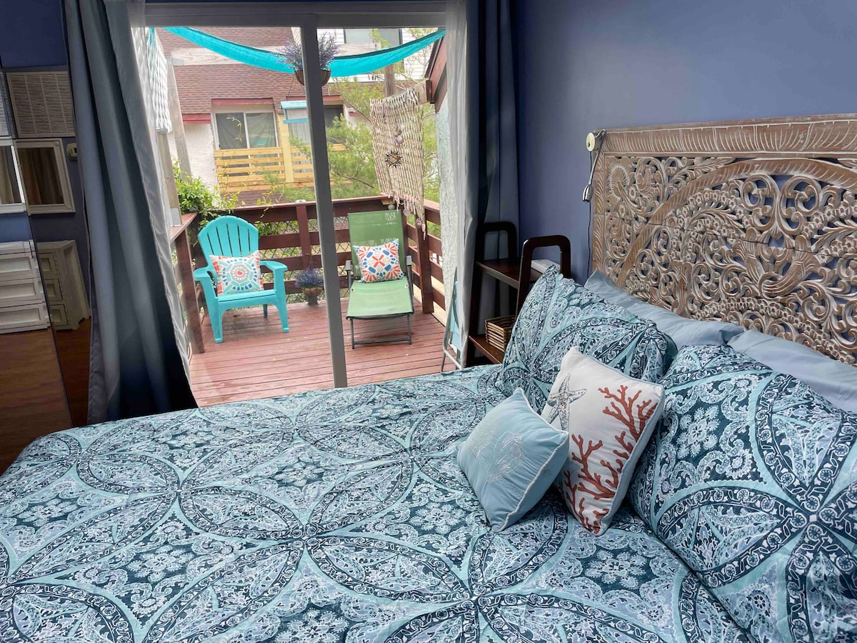 A serene bedroom features a detailed headboard and a vibrant blue-patterned bedspread. Large sliding glass doors open to a wooden deck, which is furnished with colorful chairs and a hammock, allowing natural light to fill the space.