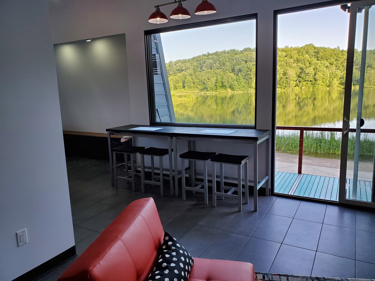 A stylish seating area features a bright red couch with black and white polka dot accents. Adjacent is a large window showcasing views of a serene lake and lush greenery. A long countertop with stools provides an inviting space for dining or conversation.