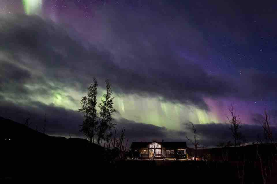 The cabin is illuminated under a night sky filled with vibrant colors from the northern lights. Soft lighting highlights the structure, creating a serene contrast with the dark landscape. Silhouettes of trees frame the scene, enhancing the tranquil atmosphere.
