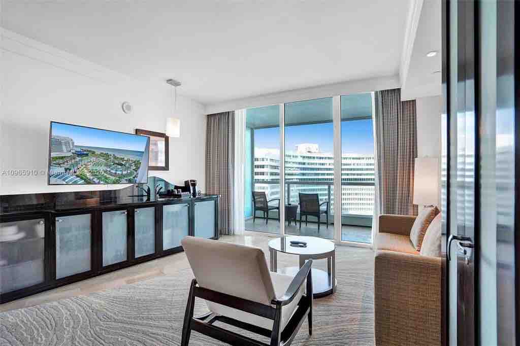 A modern living area features a fold-out sofa and a single armchair positioned around a small round table. A large flat-screen TV is mounted on a dark wood cabinet. Floor-to-ceiling sliding glass doors lead to a private balcony with views of the surrounding area.