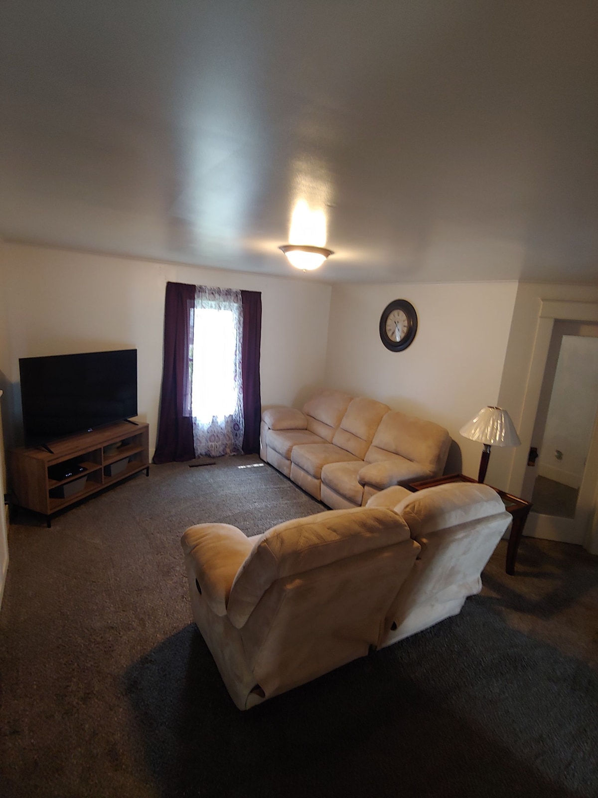 A comfortable living room is showcased with a beige sectional sofa facing a flat-screen television. A soft area rug covers the floor, and natural light enters through a window adorned with dark curtains. A small lamp and wall clock complete the inviting setting.