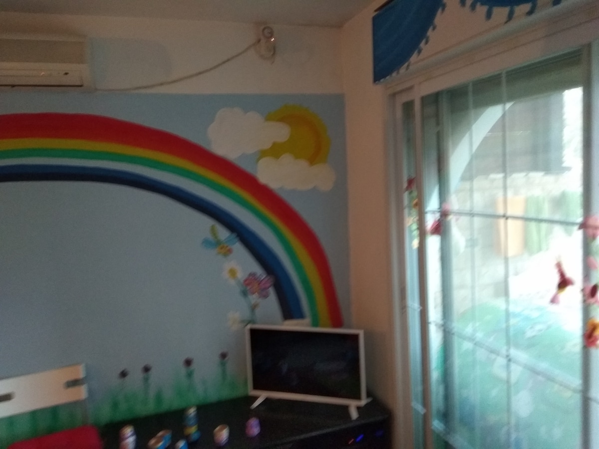 A colorful and cheerful living area is highlighted by a mural featuring a rainbow and sun against a blue sky. A small television is positioned on a dark cabinet, and a sliding glass door offers access to the outside, framed by decorative elements.