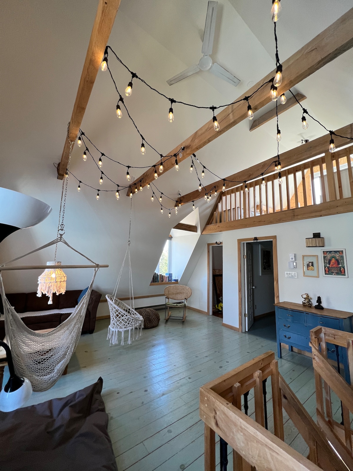 An airy living space features wooden beams and decorative string lights overhead, providing a cozy ambiance. A hammock swing and a rattan chair are positioned to invite relaxation, while a staircase adds vertical interest. A calm color palette and natural light enhance the tranquil setting.