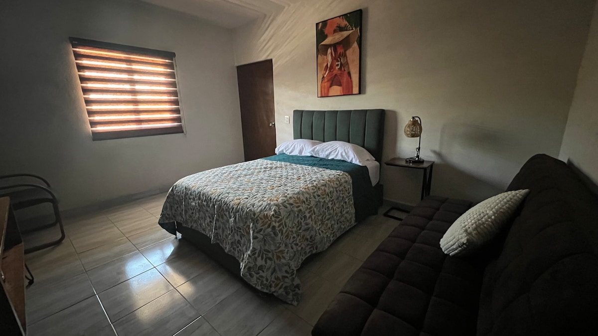 A spacious guest room features a well-made bed with a patterned blanket and a comfortable sofa alongside it. Natural light enters through a window with wooden blinds, casting soft shadows on the tiled floor. Decorative wall art adds a subtle touch of character.