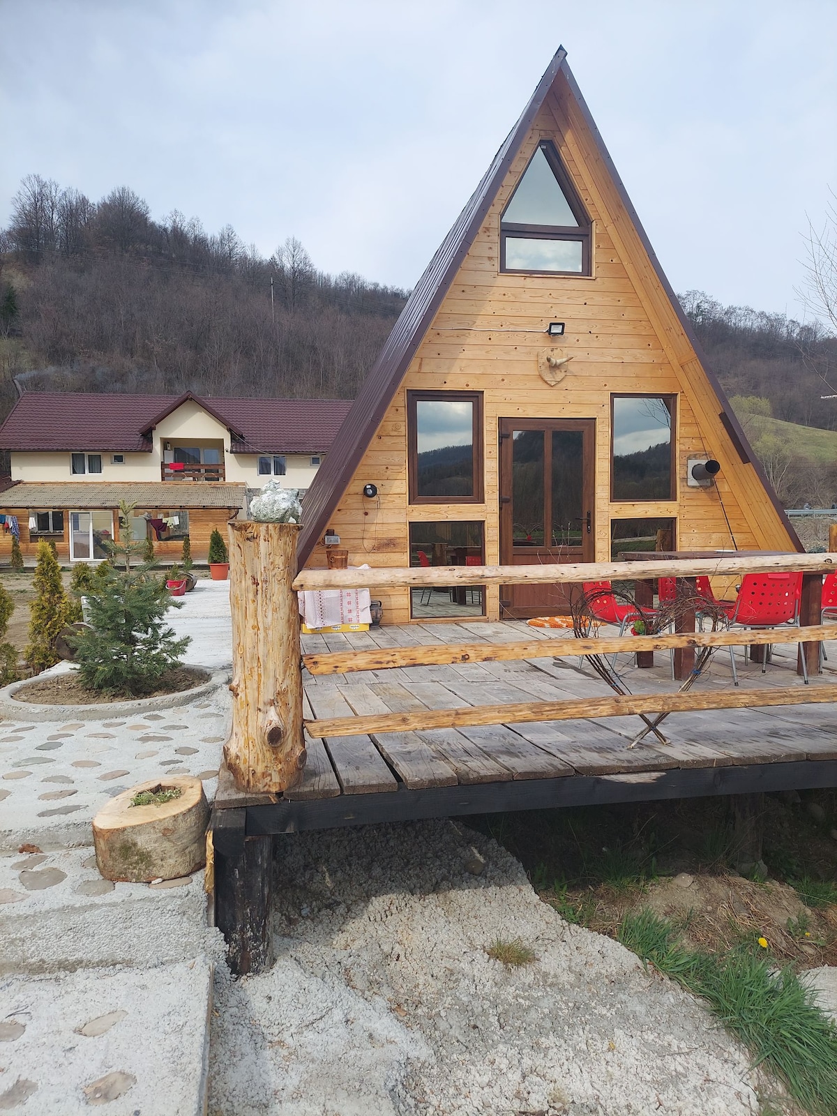 A triangular wooden cabin is situated on a stone pathway, surrounded by greenery. An outdoor deck features wooden railings and red chairs, providing an inviting seating area. In the background, another building is visible, blending harmoniously with the natural landscape.