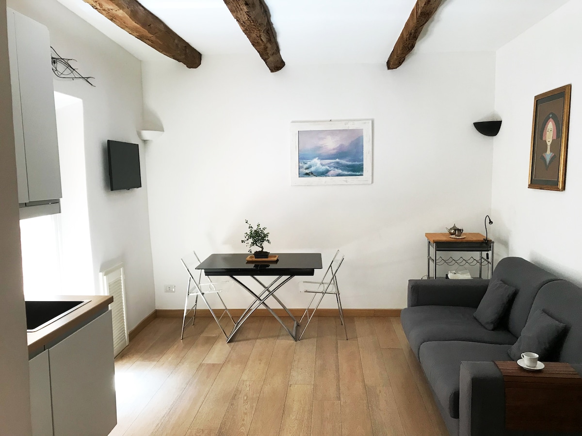 A bright living area features exposed wooden beams and light-colored walls. A gray sofa is positioned beside a small wooden table, set with a vase of greenery. A dining table with foldable chairs and a wall-mounted television are visible, adding functionality to the space.