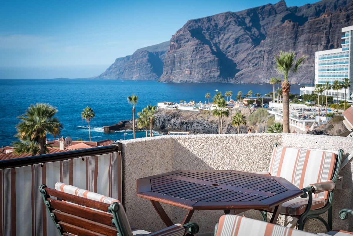 A sunlit terrace is equipped with an octagonal wooden table and matching chairs. The ocean view extends to the rugged cliffs of Los Gigantes, with palm trees lining the shore. The serene blue water contrasts with the warm hues of the surrounding landscape.