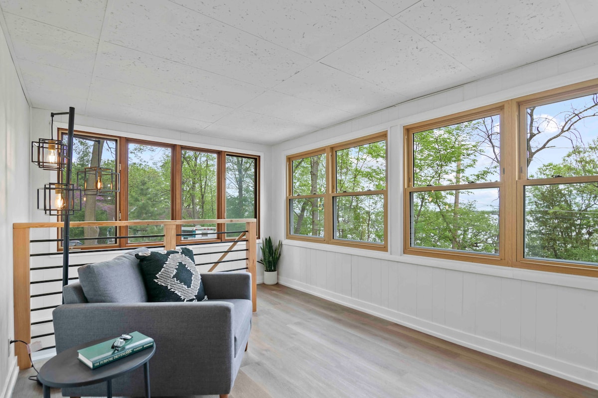 A bright sunroom is featured with large windows offering views of surrounding greenery. A cozy gray sofa with decorative cushions is positioned nearby, while a potted plant adds a touch of nature. Modern light fixtures provide illumination, enhancing the welcoming atmosphere.