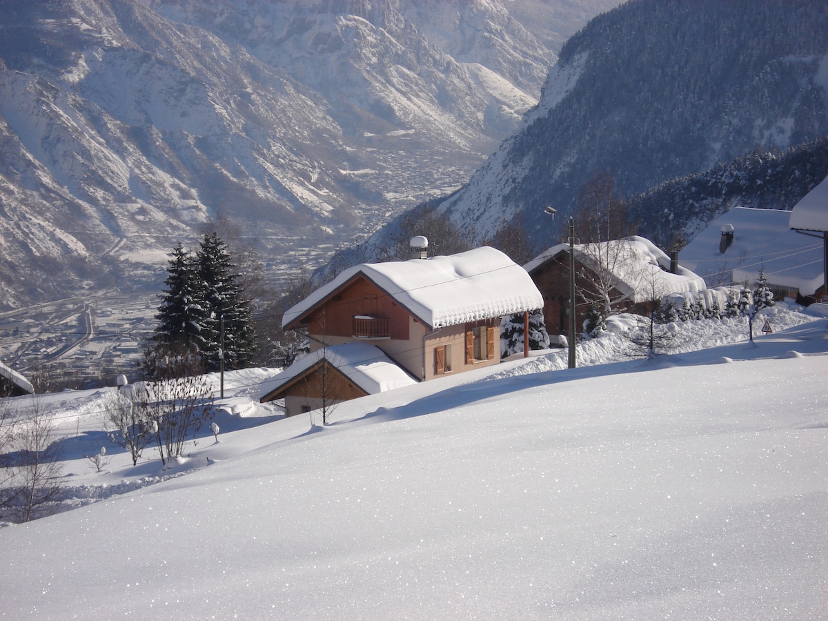 A charming mountain studio is nestled in a snowy landscape, surrounded by freshly fallen snow. The building features a sloped roof covered in snow, with trees and neighboring homes visible in the background. Majestic mountains rise beyond, creating a serene winter scene.