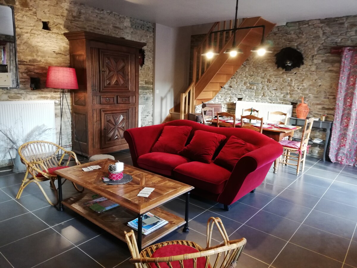 A cozy living area is shown with a bright red sofa and a wooden coffee table. Warm lighting highlights a rustic stone wall and wooden staircase in the background. Rattan chairs add to the inviting arrangement, which includes a dining space with wooden tables and colorful decor.