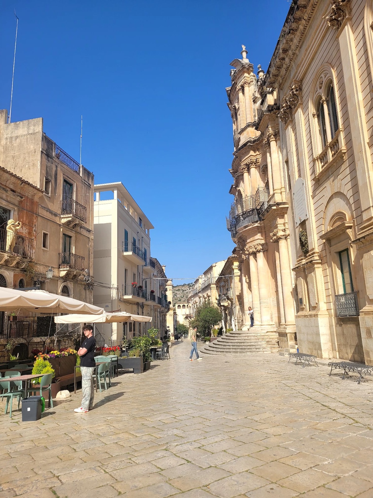 A charming street view is presented, featuring a sunlit plaza lined with quaint cafes and historic buildings. Stone-paved paths lead past vibrant outdoor seating, with elements of Baroque architecture prominently displayed on one side. Clear blue skies create a bright atmosphere.