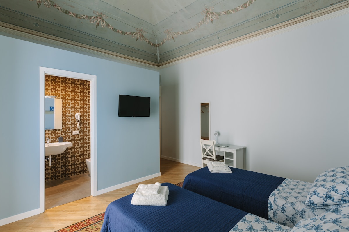 A well-lit bedroom features two twin beds with blue covers, complemented by decorative pillows. An inviting en-suite bathroom is visible through an open door. A flat-screen television is mounted on the wall, and a simple desk and chair sit against one side. Decorative wall patterns add character.