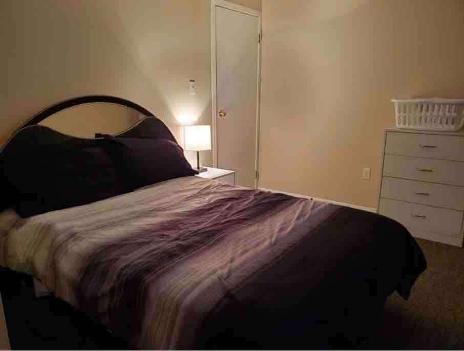 A cozy bedroom features a spacious bed with dark bedding accented by subtle stripes. A modern nightstand hosts a lamp providing soft light. A white laundry basket is placed beside a light-colored dresser, and a closed door can be seen in the background.