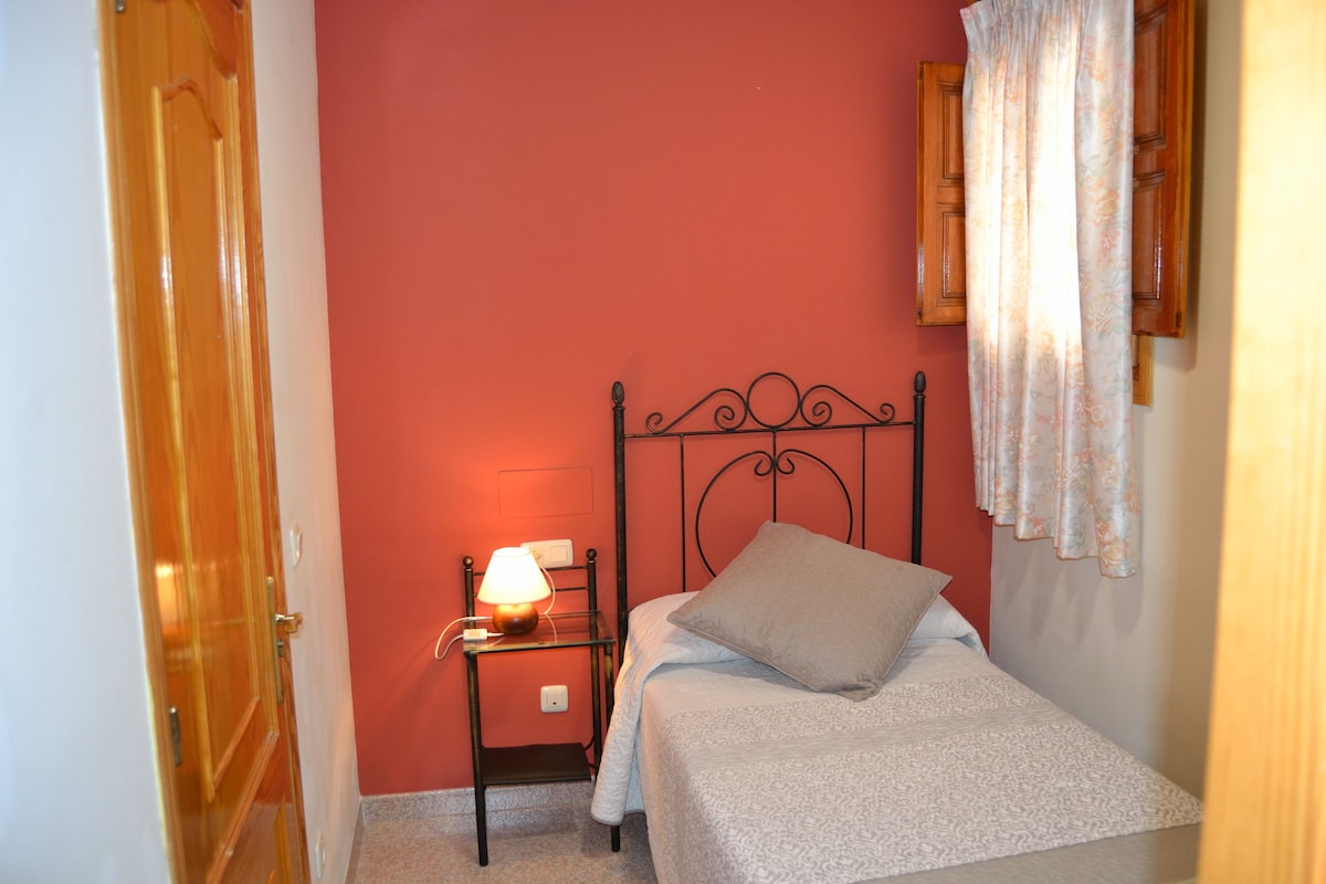 A single bedroom features a cozy bed with decorative pillows, accompanied by a bedside table with a lamp. Natural light enters through a window adorned with light curtains, while the warm red wall adds an inviting touch to the space.