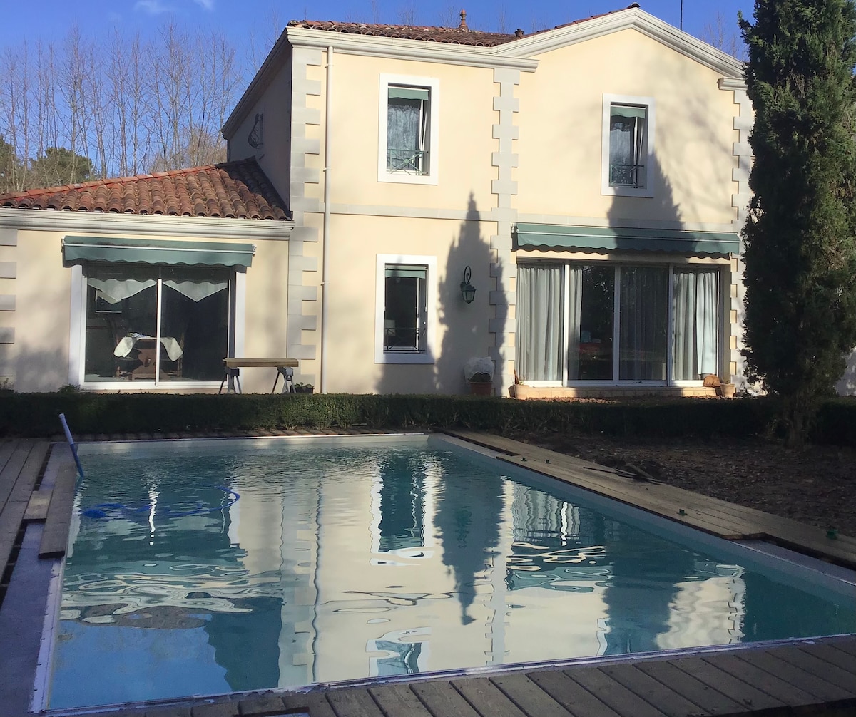 The exterior of a charming two-story house is visible, featuring large windows that reflect the sunlight. A swimming pool is situated in front of the house, surrounded by wooden decking. Lush greenery is present, enhancing the tranquil ambiance of the property.