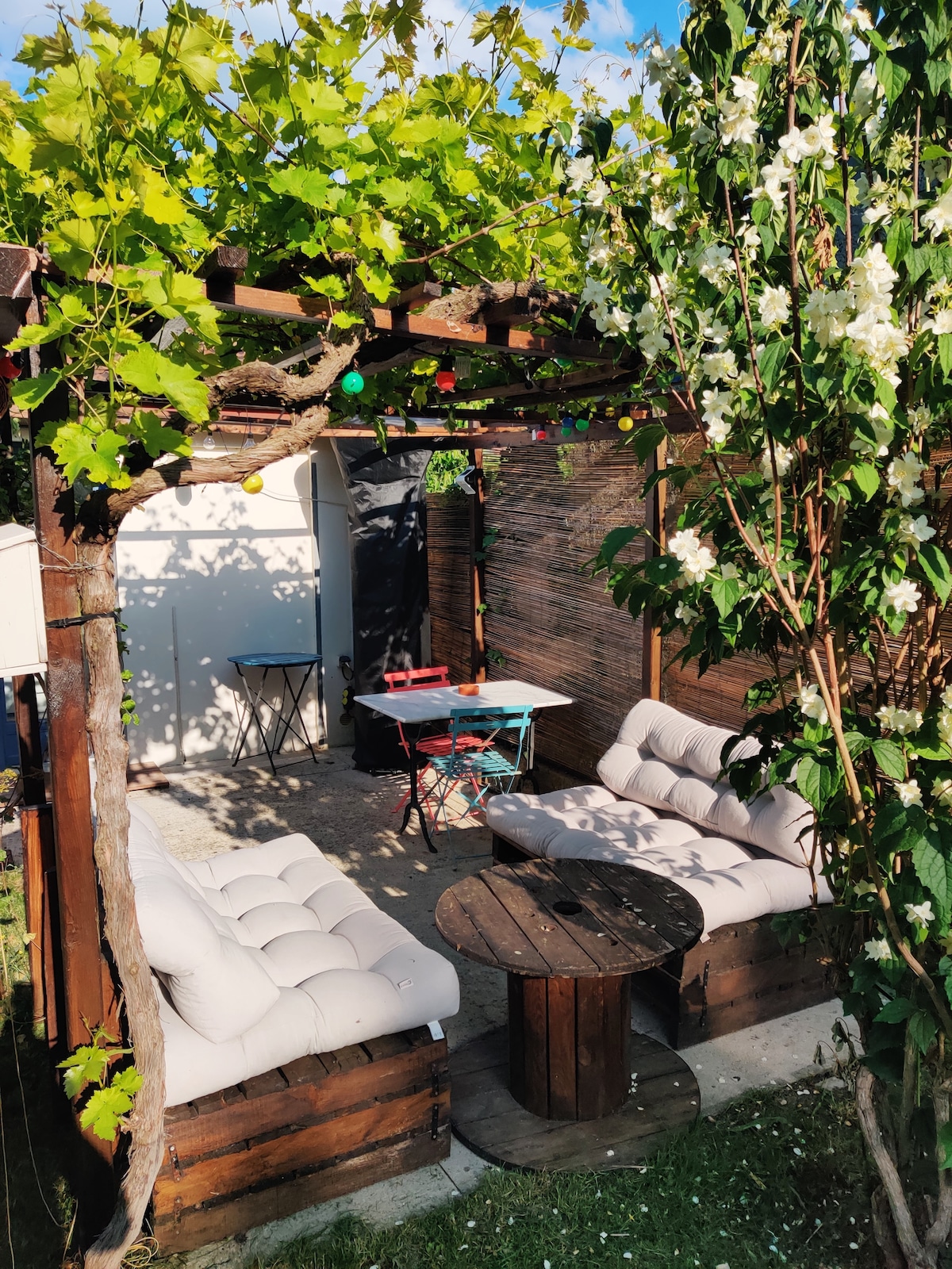 An outdoor seating area is nestled under a vine-covered pergola, featuring soft, light-colored seating arranged around a circular wooden table. Decorative lights and greenery enhance the space, providing a serene environment for relaxation or dining.