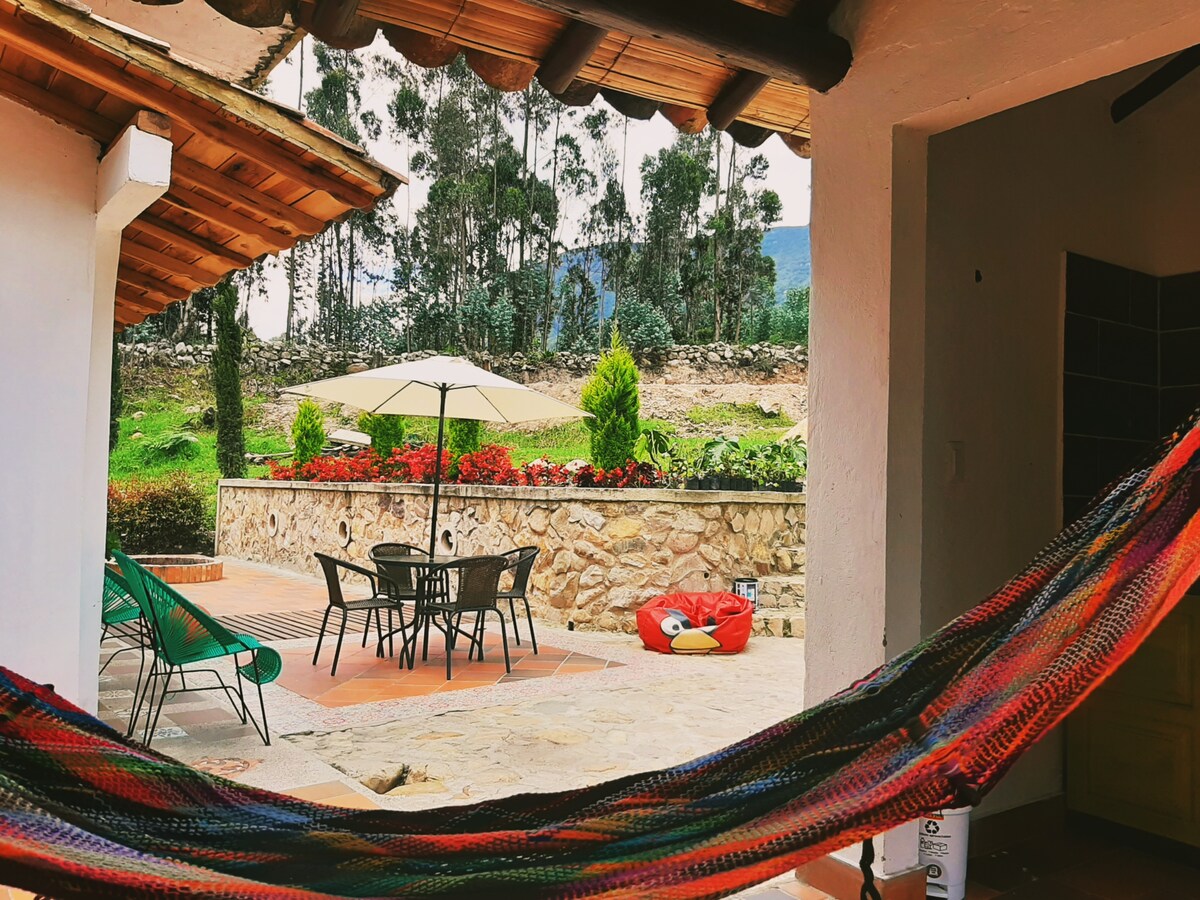 A colorful hammock is suspended between a wooden structure and a support post. Beyond, a patio area is visible with a circular table and black chairs under a large umbrella. A garden adorned with flowering plants and a stone wall provides a natural backdrop.