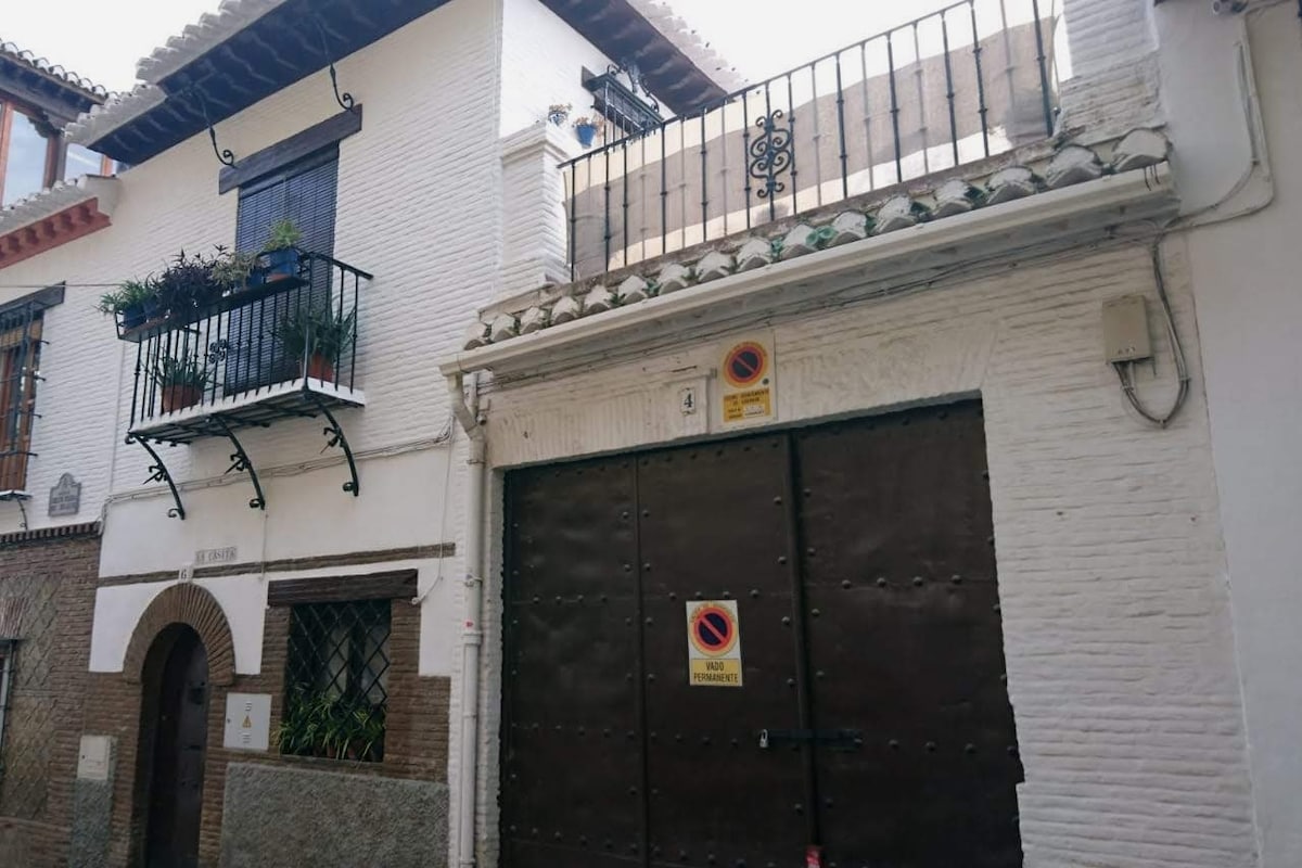 The exterior of a two-story white casita features a traditional Spanish design, with decorative wrought iron balconies adorned with potted plants. A large wooden door provides access to the main entry, while a roof terrace area is visible above, framed by a decorative railing.