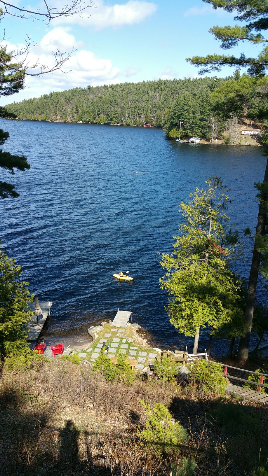 A serene view of a calm lake surrounded by lush greenery, with small pine trees framing the scene. A canoe glides across the water towards two docks, amidst the gentle ripples of the lake. Stone pathways lead down to the water's edge, inviting exploration.