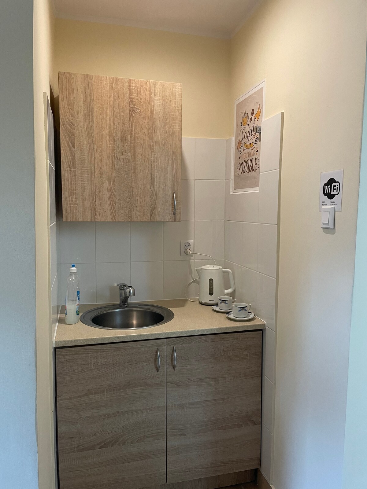 A compact kitchenette is featured, showcasing wooden cabinetry and a sink. A kettle is positioned beside stacked cups on the countertop. A small board on the wall displays a motivational print, while the space is brightened by natural light.