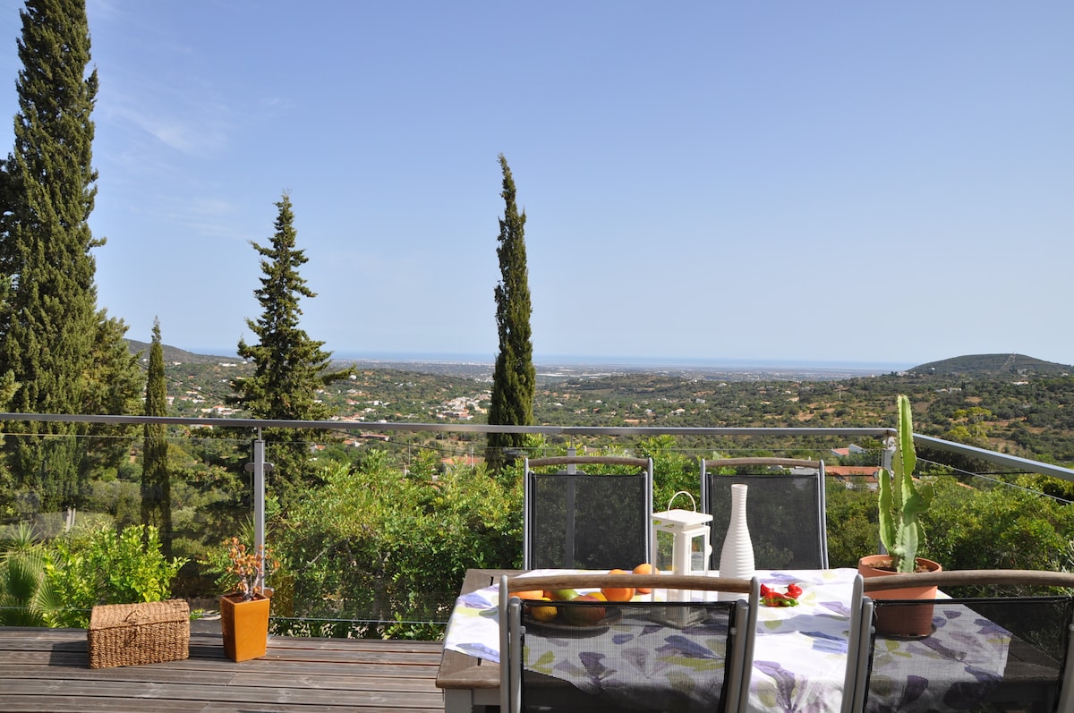 A spacious terrace offers a dining table set for outdoor meals, surrounded by lush greenery and overlooking a scenic landscape. The ocean is visible in the distance, framed by tall pine trees. Sunlight brightens the area, creating a serene atmosphere.