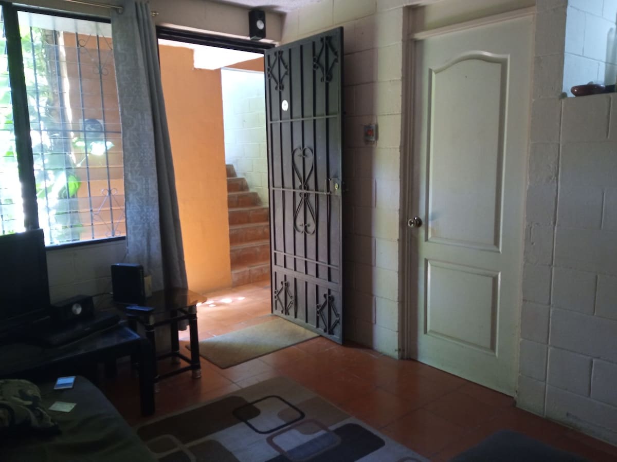 A doorway opens into a bright space, revealing terracotta tiled floors. Natural light flows through a large window, while a comfortable seating area is present. A dark metal door with decorative patterns adds character, and stairway access can be seen leading to an upper level.