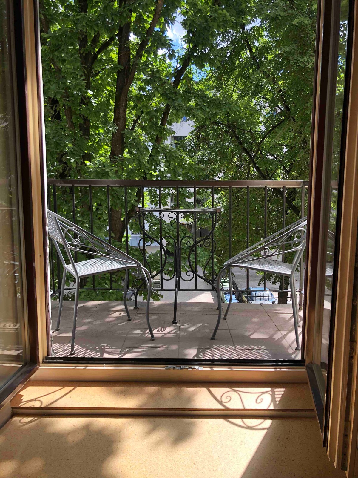 A small balcony is seen through open glass doors, featuring a round table and two metal chairs. Lush greenery from nearby trees provides natural shade and tranquility, enhancing the outdoor space's inviting atmosphere.