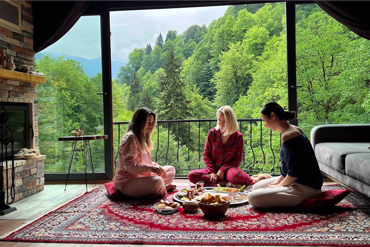 A spacious living area features large windows that frame a lush green view of surrounding trees. A traditional patterned rug is placed on the floor, with three individuals sitting on it, enjoying a meal arranged on a low table. A stone fireplace adds a cozy touch to the atmosphere.