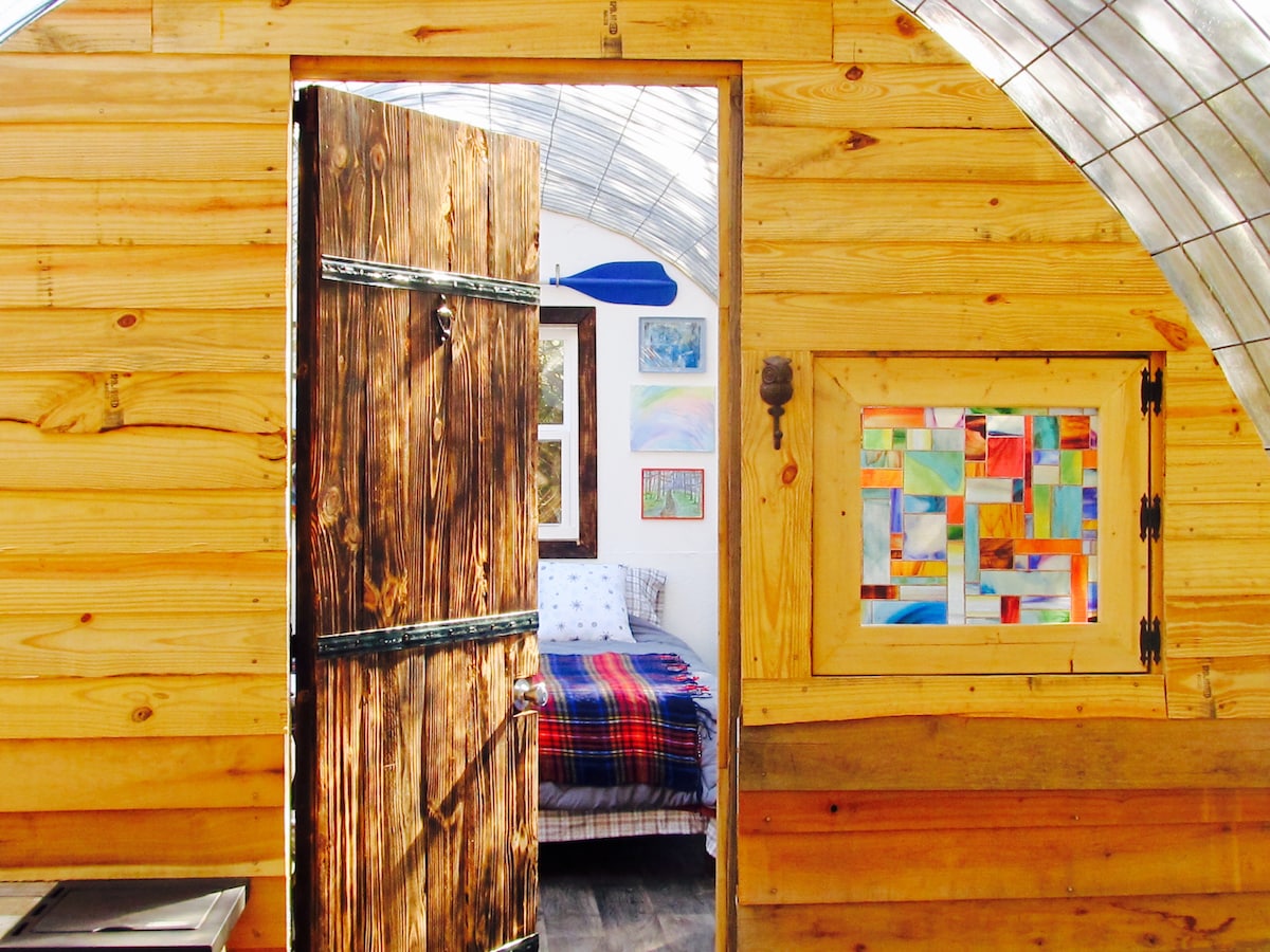 The entrance to the Stargazing hut features a wooden door and a stained glass window, allowing natural light to enter. Inside, a cozy sleeping area is visible, adorned with colorful artwork and a plaid blanket on the bed, creating a warm and inviting space.