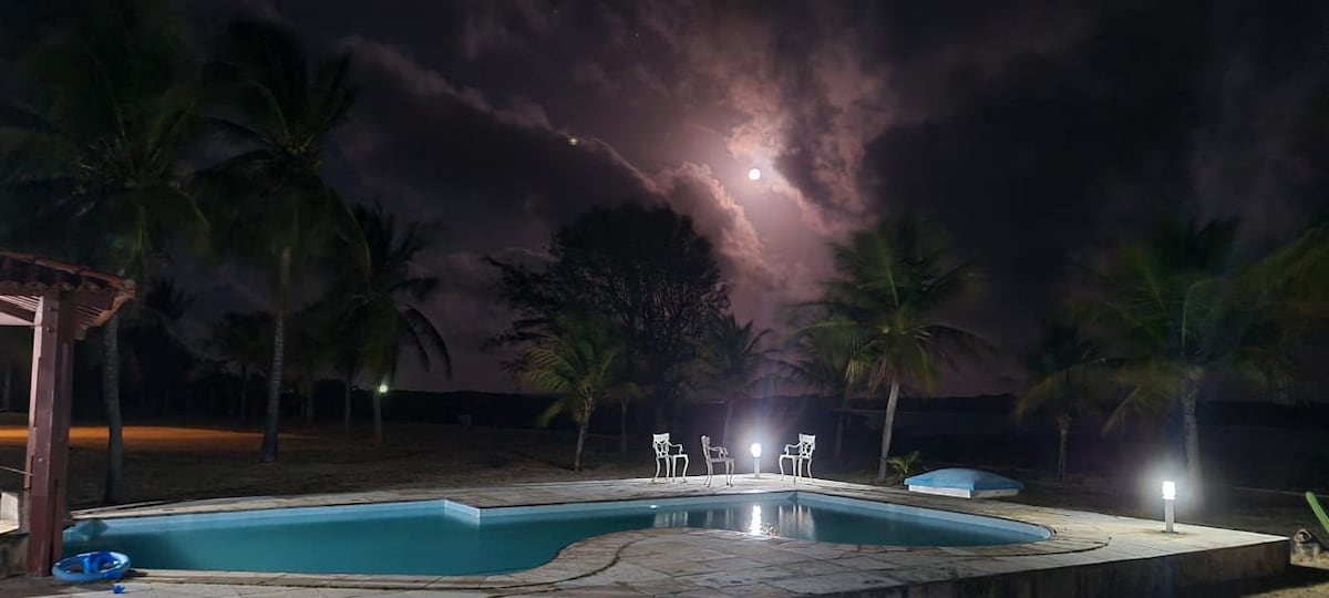 A tranquil swimming pool is illuminated under the night sky, surrounded by palm trees. The moon reflects on the water's surface, creating a serene atmosphere. Nearby, casual seating is visible, enhancing the outdoor gathering space.