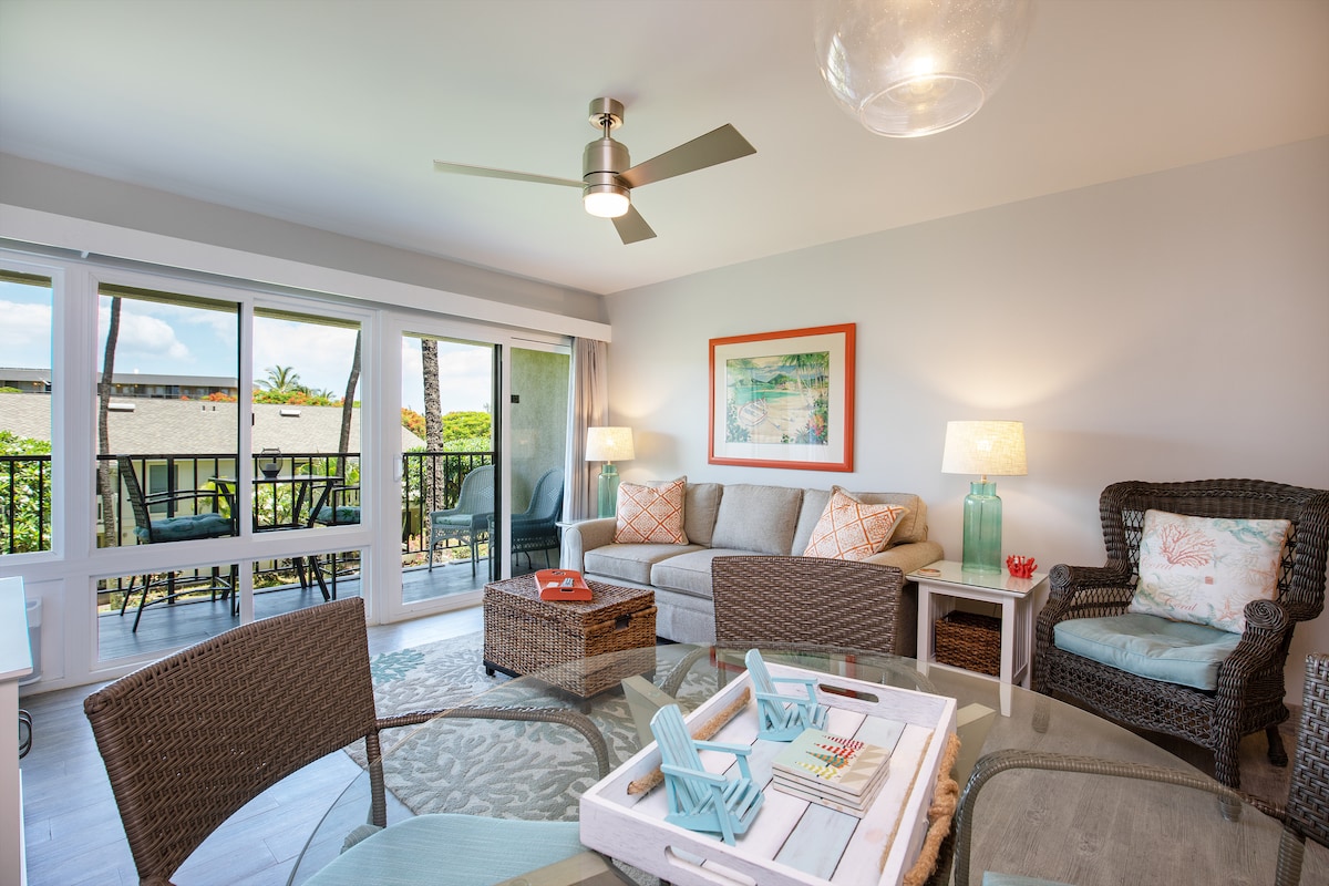 A bright living area features a spacious sofa and wicker chairs, accompanied by a decorative coffee table with beach-themed accents. Natural light flows in through large sliding doors, revealing a view of greenery outside. Two lamps provide soft illumination, enhancing the room's inviting ambiance.