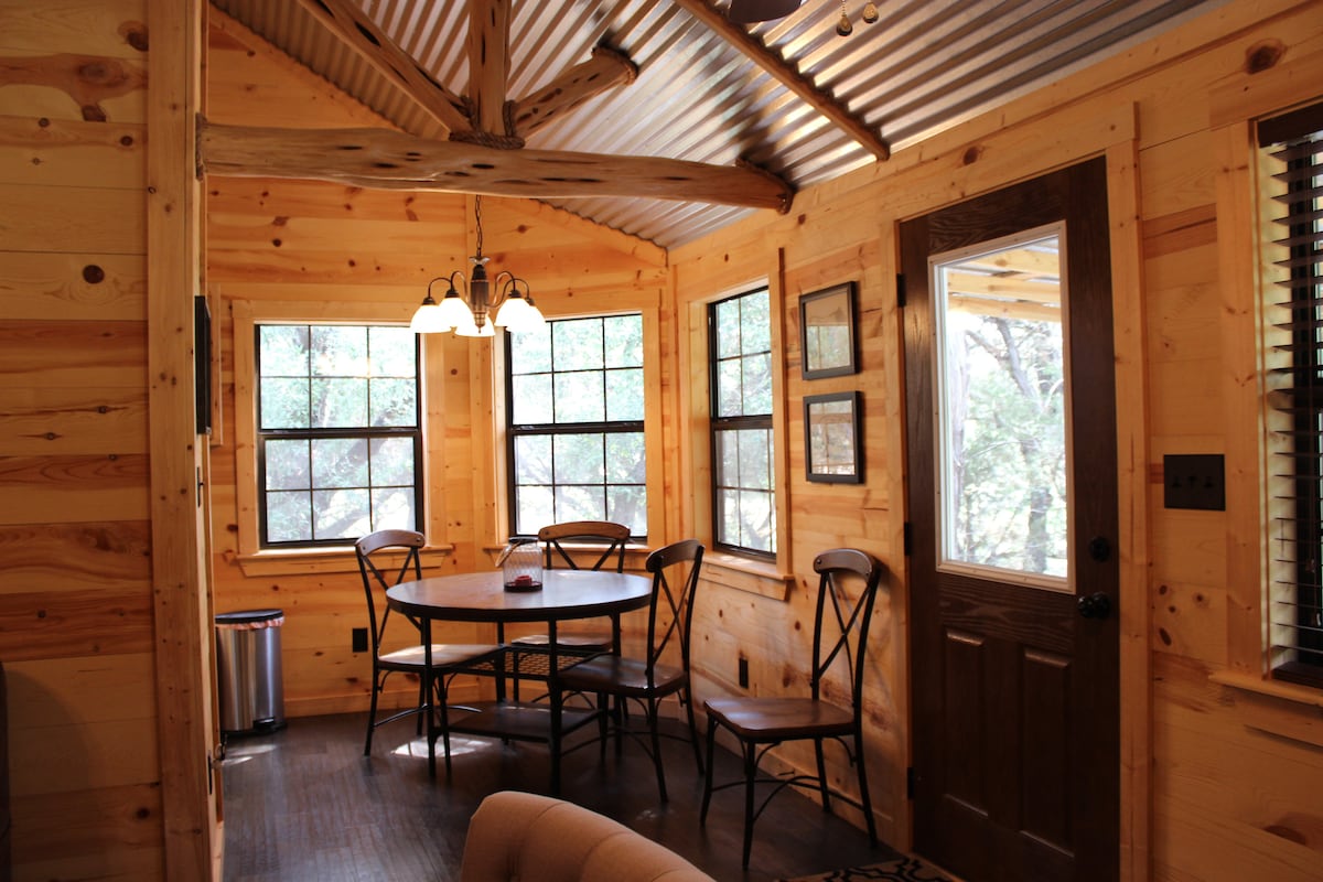 A rustic dining area features a round wooden table surrounded by four chairs. Large windows allow natural light to fill the space, showcasing the wooden walls and beams. A door leads outside, and a waste bin is visible in the corner.