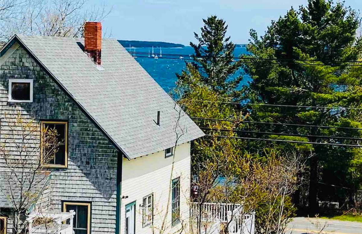 A view of a traditional cottage set against a backdrop of trees and the ocean. The cottage features a grey shingle roof and light-colored siding. Beyond the house, sailboats can be seen on the water, framed by the greenery of the surrounding landscape.