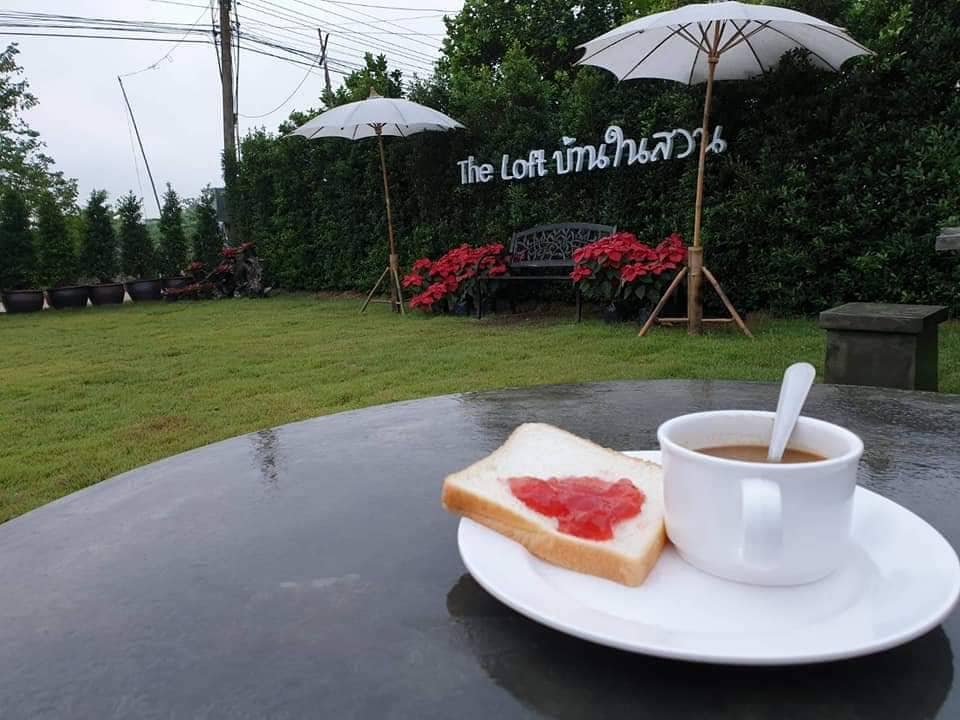 A table set with a white plate holding a slice of bread topped with red jam and a cup of coffee. In the background, two white parasols provide shade over vibrant greenery, with a sign reading 'The Loft' visible among neatly arranged flowers.