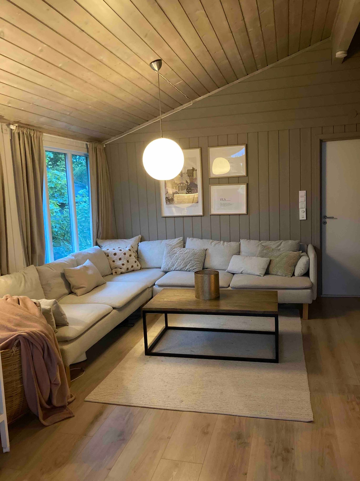 A comfortable living area features a spacious sectional sofa adorned with various cushions. A sleek coffee table is centered on a light-colored rug, while soft natural light filters through large windows, enhancing the warm wood paneling of the walls.