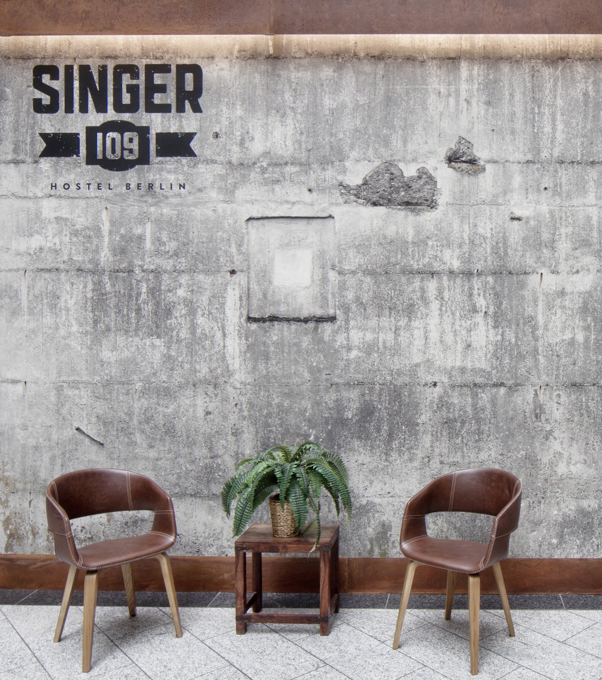 Two brown chairs are positioned beside a small wooden table, topped with a green potted plant. A textured, grey concrete wall serves as the backdrop, featuring the Singer 109 Hostel logo prominently displayed at the top.