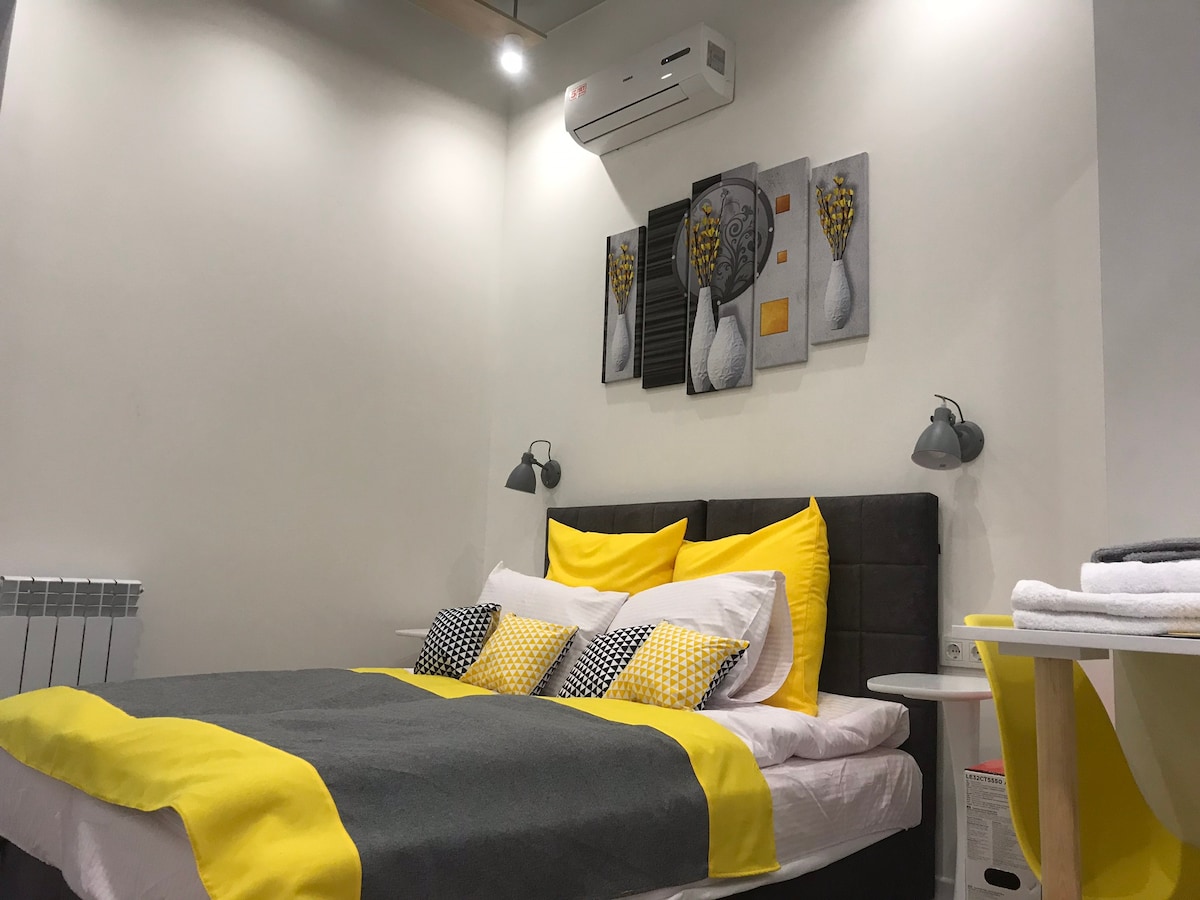 A cozy bedroom features a spacious bed adorned with yellow and gray bedding. Above the bed, modern artwork depicting floral elements hangs on the wall. Wall-mounted lamps are positioned on either side, and a small table is visible nearby.