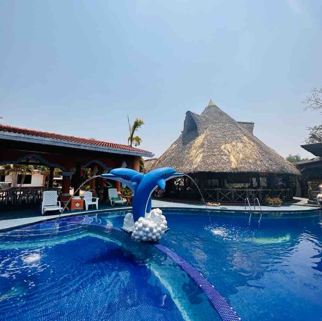 A vibrant pool area is featured, with a playful dolphin fountain creating a water spray. Surrounding the pool, lounge chairs are arranged for relaxation, and a traditional thatched-roof building provides shade and dining options. Clear blue skies enhance the outdoor atmosphere.