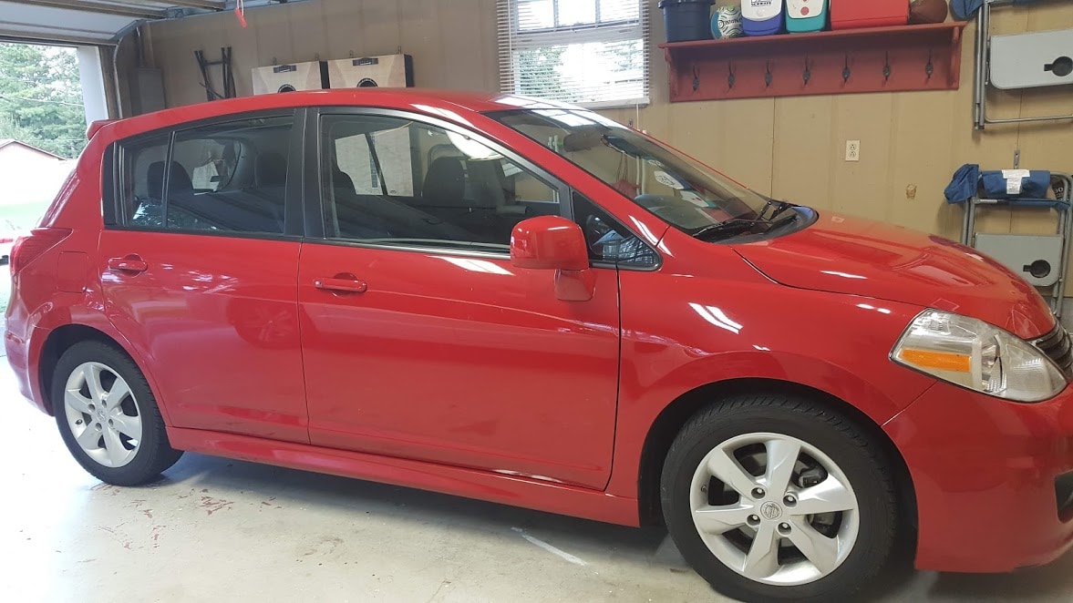 A red compact car is parked in a garage space, showcasing its sleek exterior and clean lines. The vehicle features five doors and is positioned adjacent to a wall with storage shelves arranged above and tools visible in the background.