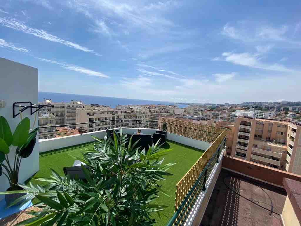 A spacious rooftop terrace features a green lawn with seating areas, offering panoramic views of the sea and city. Lush plants add a touch of greenery against a backdrop of bright blue sky and distant buildings.