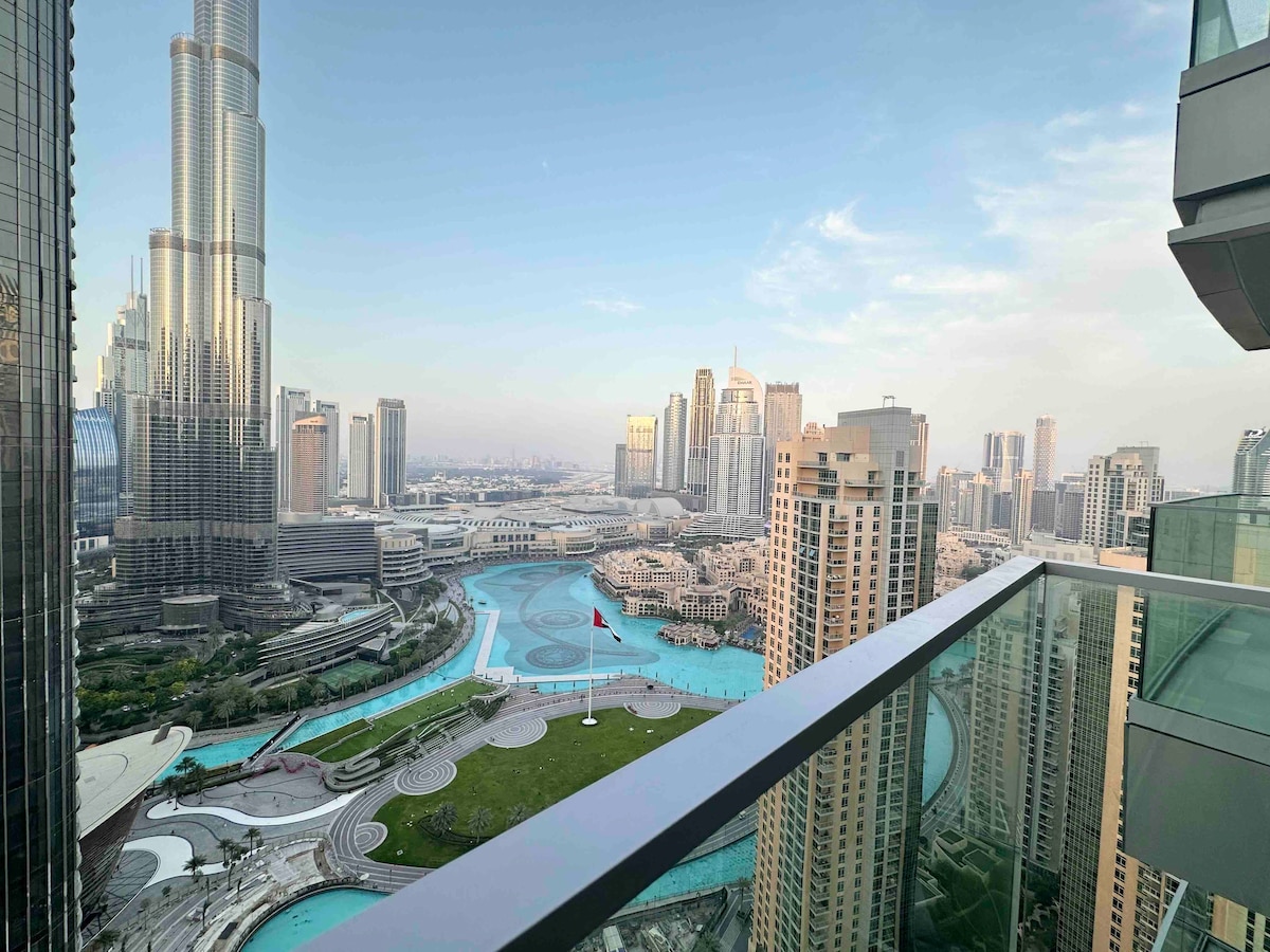 A panoramic view is displayed from the balcony, showcasing the iconic Burj Khalifa alongside pristine city scenery. The Dubai Fountain can be seen below, with its colorful water displays. High-rise buildings frame the skyline, creating a vibrant urban landscape against a clear sky.
