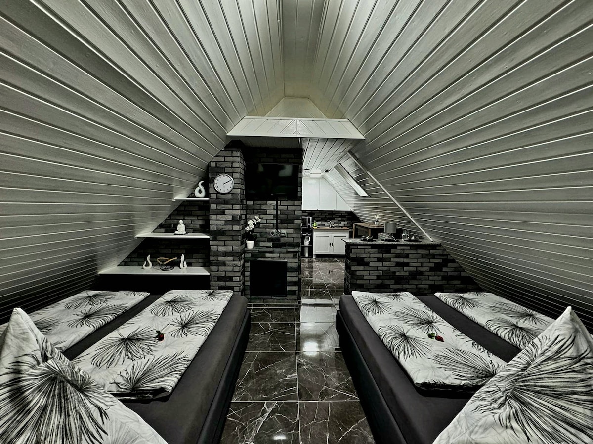 A spacious attic room features two large double beds with patterned bedding and decorative pillows. Sleek dark tiles cover the floor, and modern shelving is mounted on the slanted walls. A functional kitchen area with appliances is visible in the background, complemented by stylish lighting.