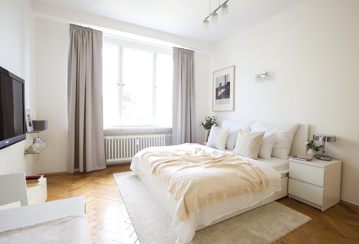 A bright bedroom features a spacious bed adorned with neutral linens and several pillows. Natural light fills the room through large windows, framed by soft curtains. A television is positioned near the bed, while a small nightstand holds a lamp and decorative elements.